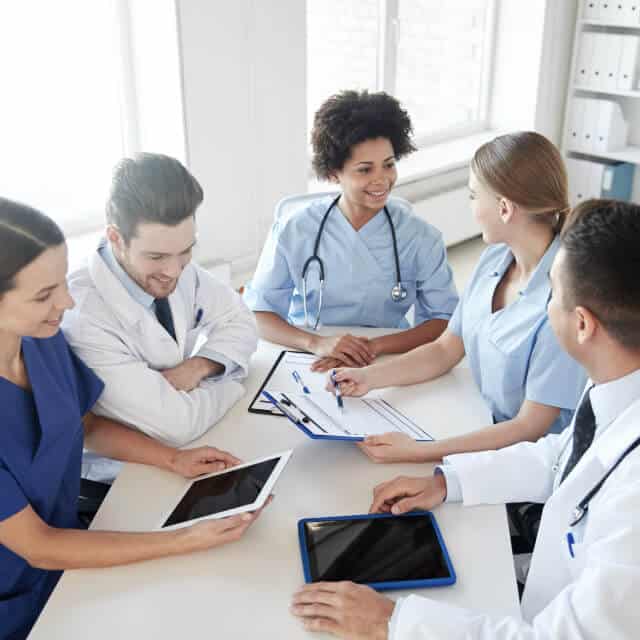 A group of doctors of various sexes and ethnicities have a conversation with clipboards and tablets in hand.