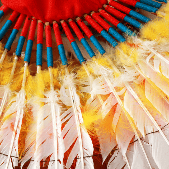 Colourful feathers with red and blue string wrapped around the ends of the feathers that are then attached in a circular shape to a red piece of fabric