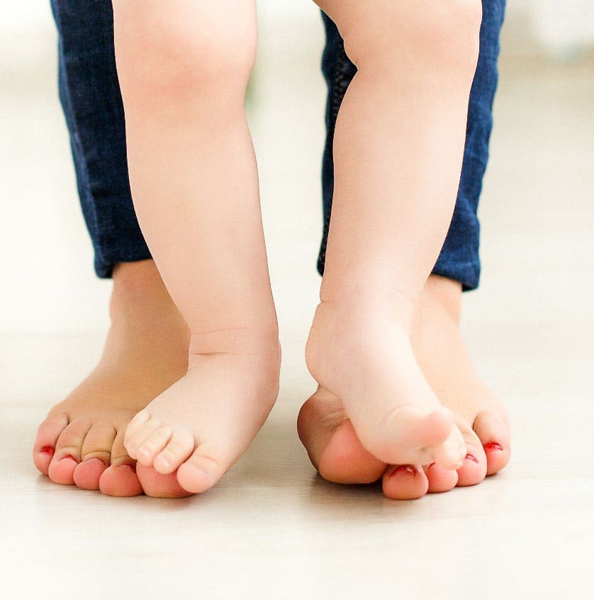 The bare feet of a baby standing on top of a female adult's bare feet