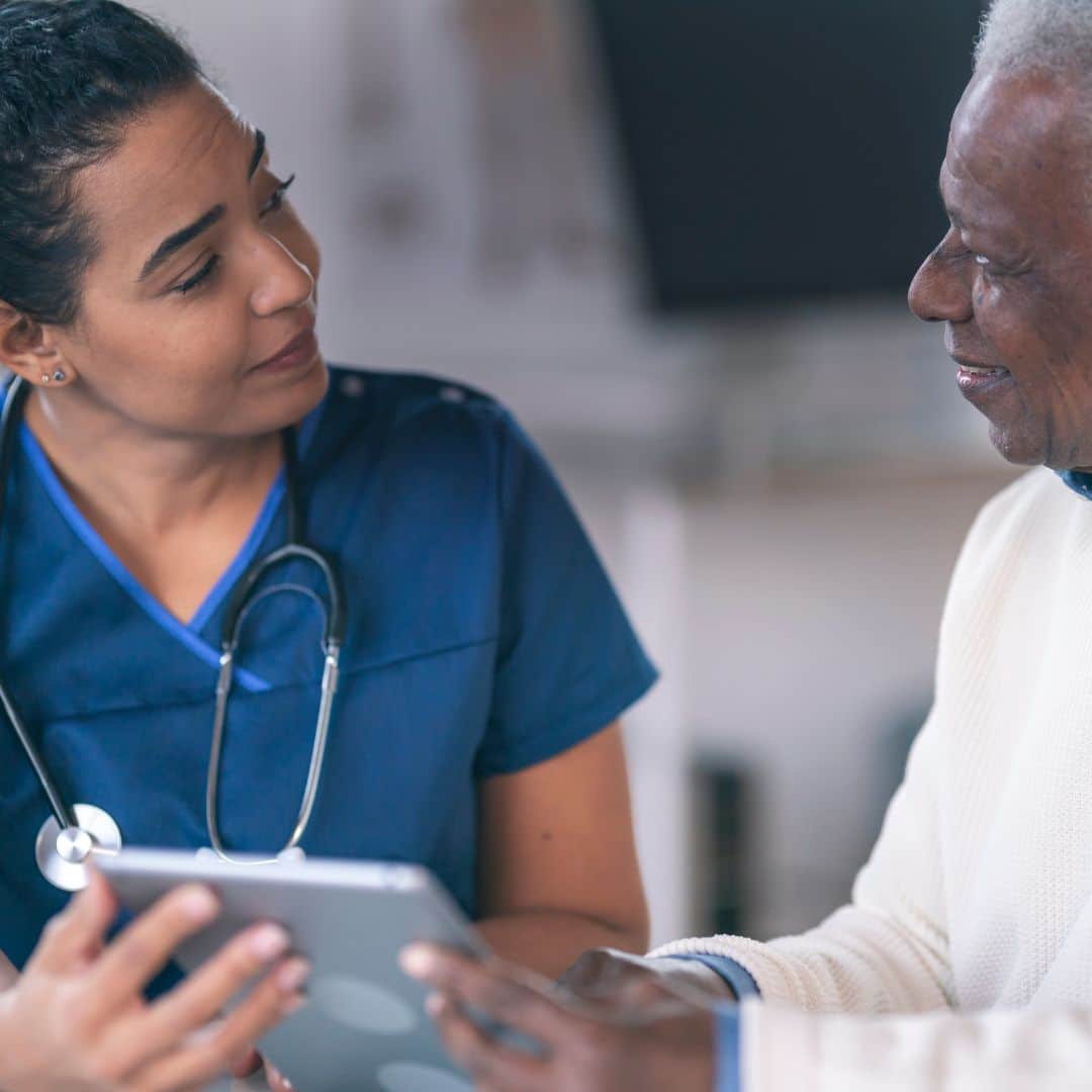 Female healthcare provider wearing a stethoscope and speaking with an older gentleman while they both hold a tablet together