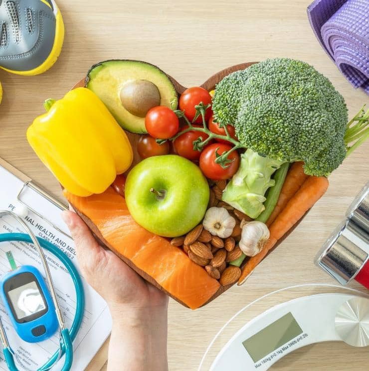 Fruit, vegetables, and salmon in a heart shaped bowl, stethoscope, blood sugar monitor, and blood sugar tracking sheet on a clipboard, exercise equipment and scale