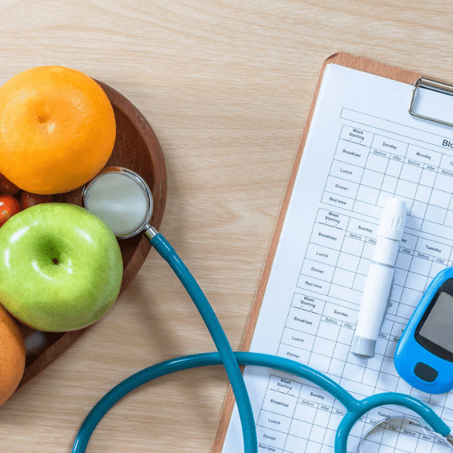 Fruit in a bowl, stethoscope, blood sugar monitor, and blood sugar tracking sheet on a clipboard