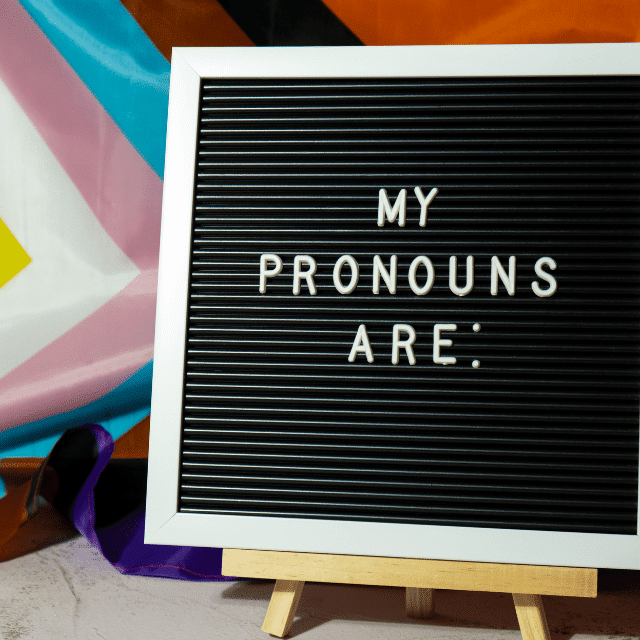 Sign that reads "My pronouns are" in front of a progressive pride flag