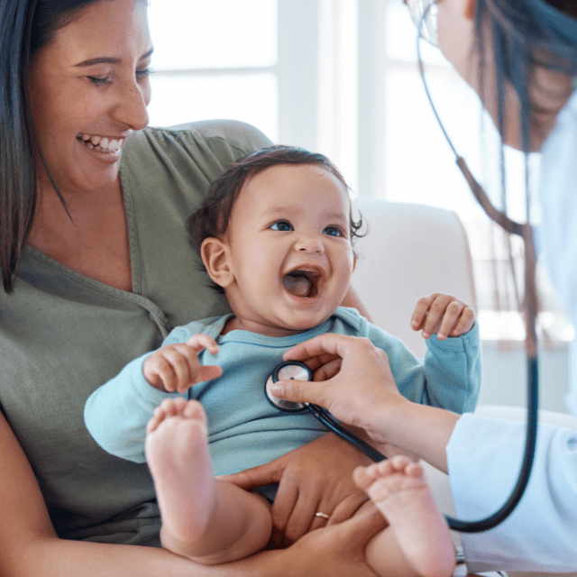 Healthcare provider using a stethoscope on a smiling baby while they are held in their mother's arms