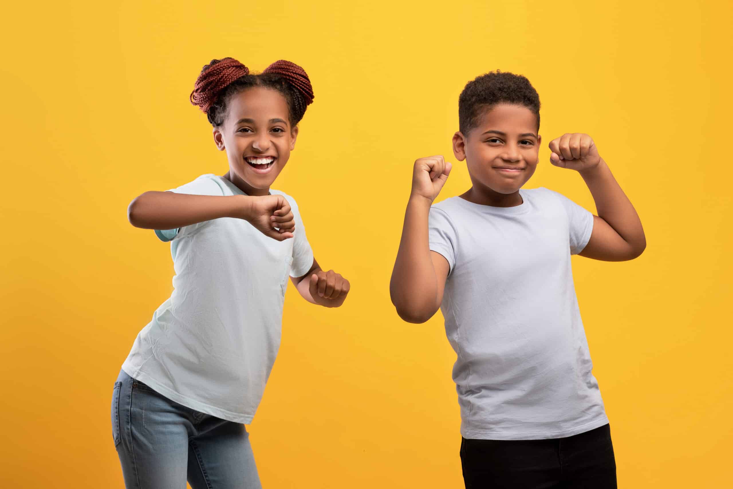 Two positive black children dancing together and cheerfully smiling over yellow studio background, adorable african american brother and sister having fun together, wearing white t-shirts