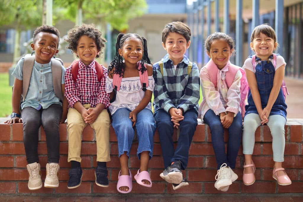 Portrait Of Multi-Cultural Elementary School Pupils Sitting On Wall Outdoors At School.