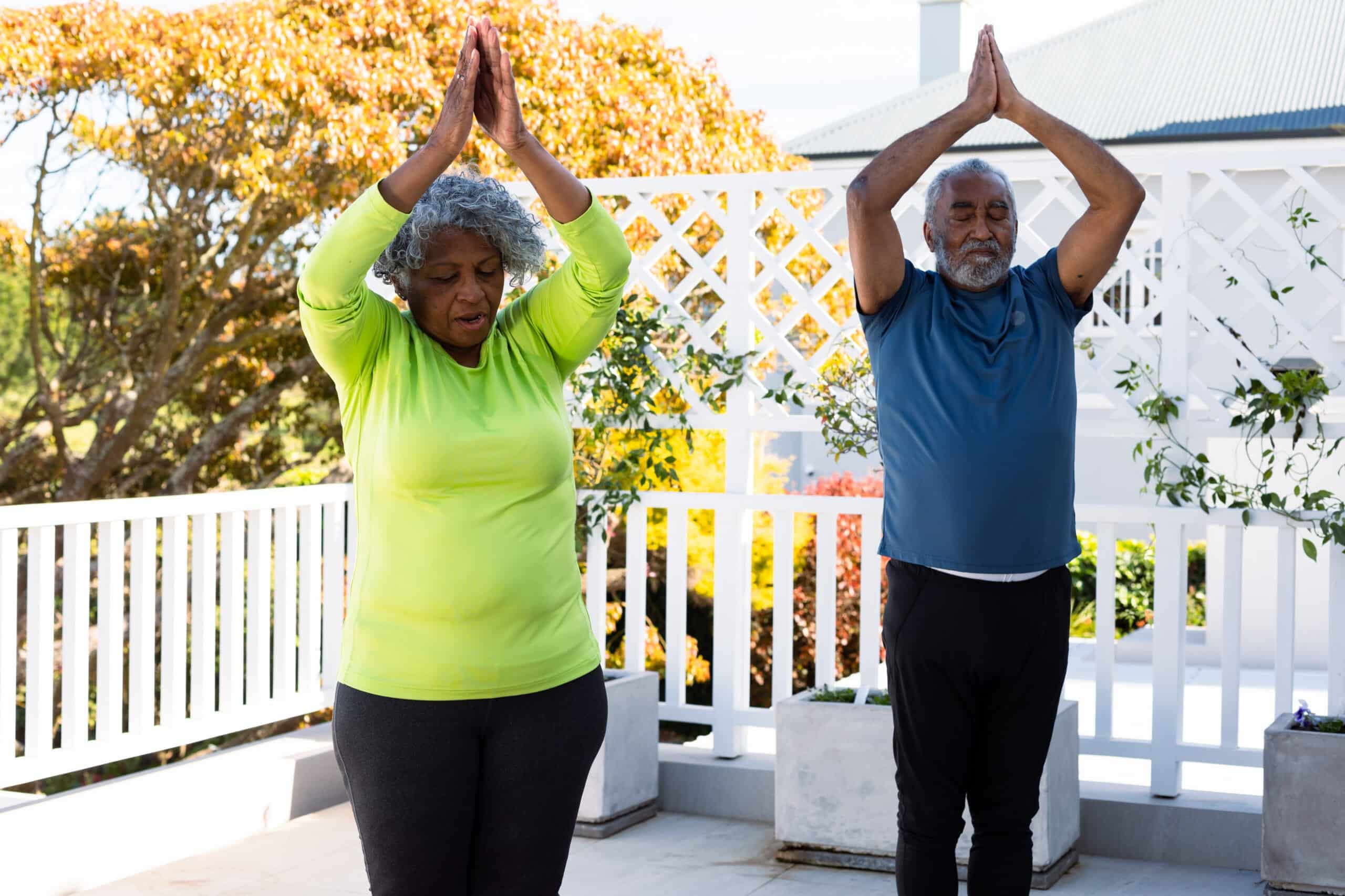 A mature woman with short, curly grey hair wearing a bright green top and dark grey pants does yoga alongside a bearded senior man in a blue top and black pants. They both have their hands raised in a praying position, with arms overhead. They stand on a patio with a white lattice fence behind them. There is foliage and colorful trees visible in the background. This peaceful photo would be ideal for projects related to health, fitness, relationships, retirement, and active lifestyle