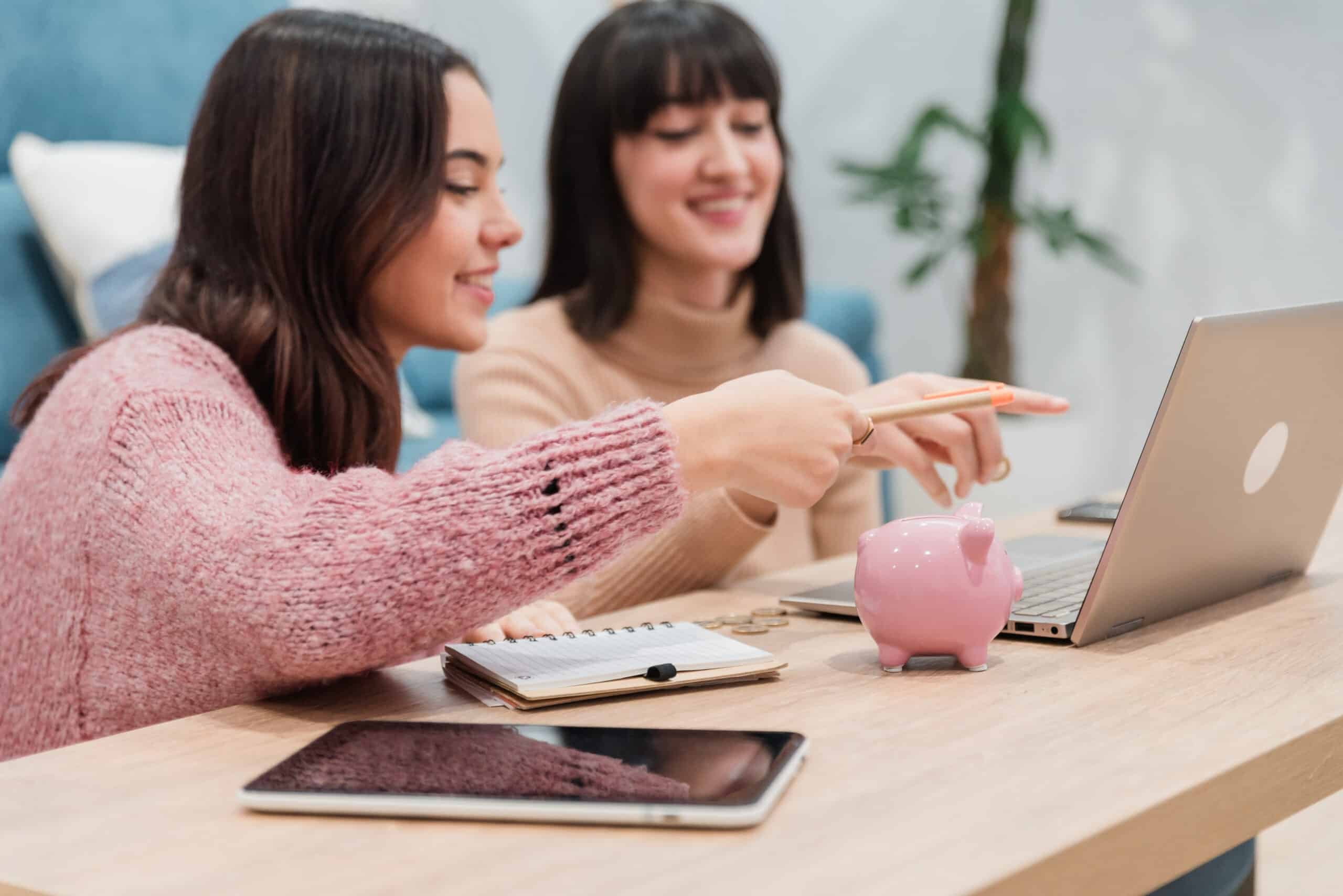 Positive young female friends sitting near sofa in living room together and using laptop while smiling happily