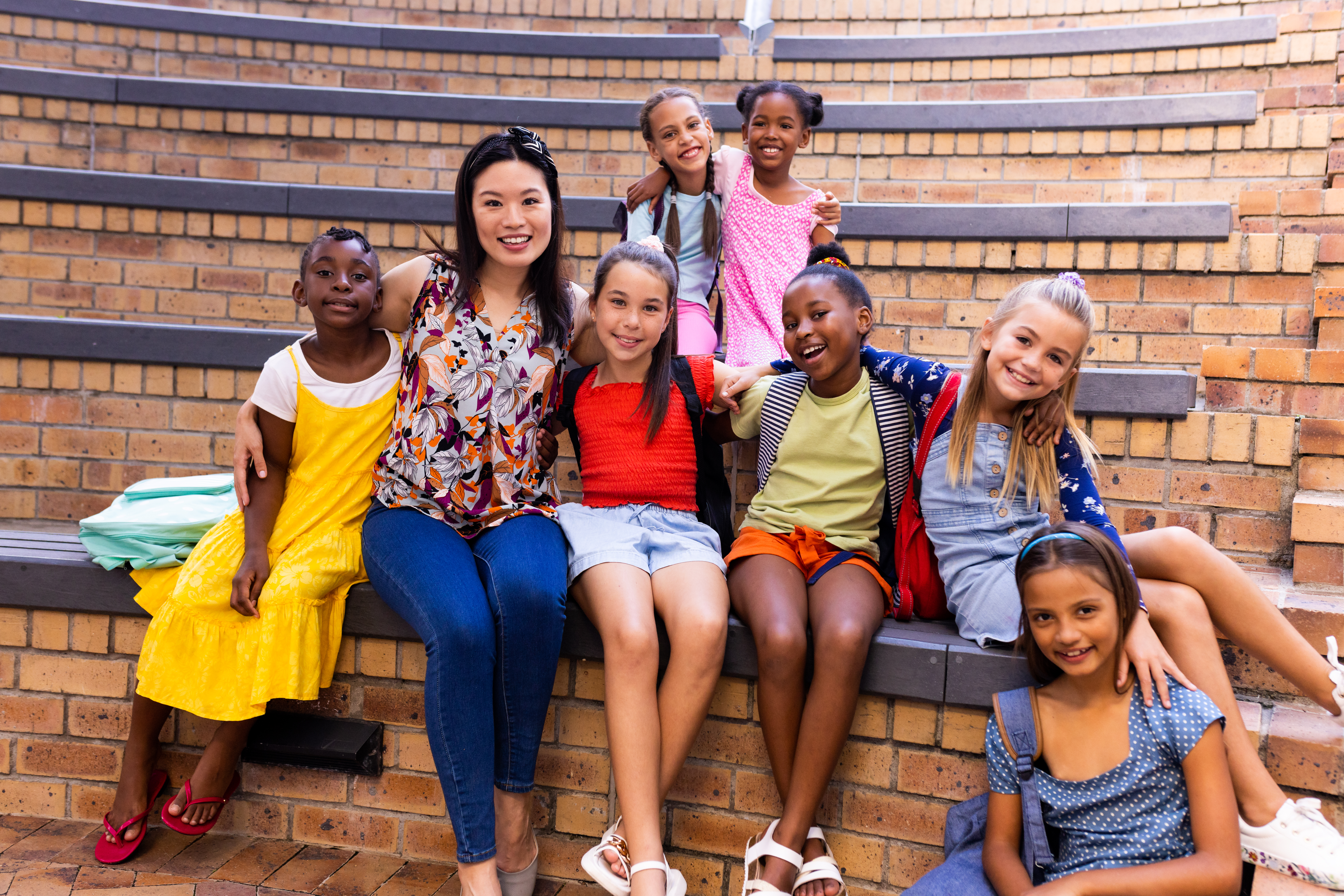 Portrait of diverse female teacher and schoolgirls sitting in elementary school outdoor auditorium. School, learning, childhood, teaching and education, unaltered.