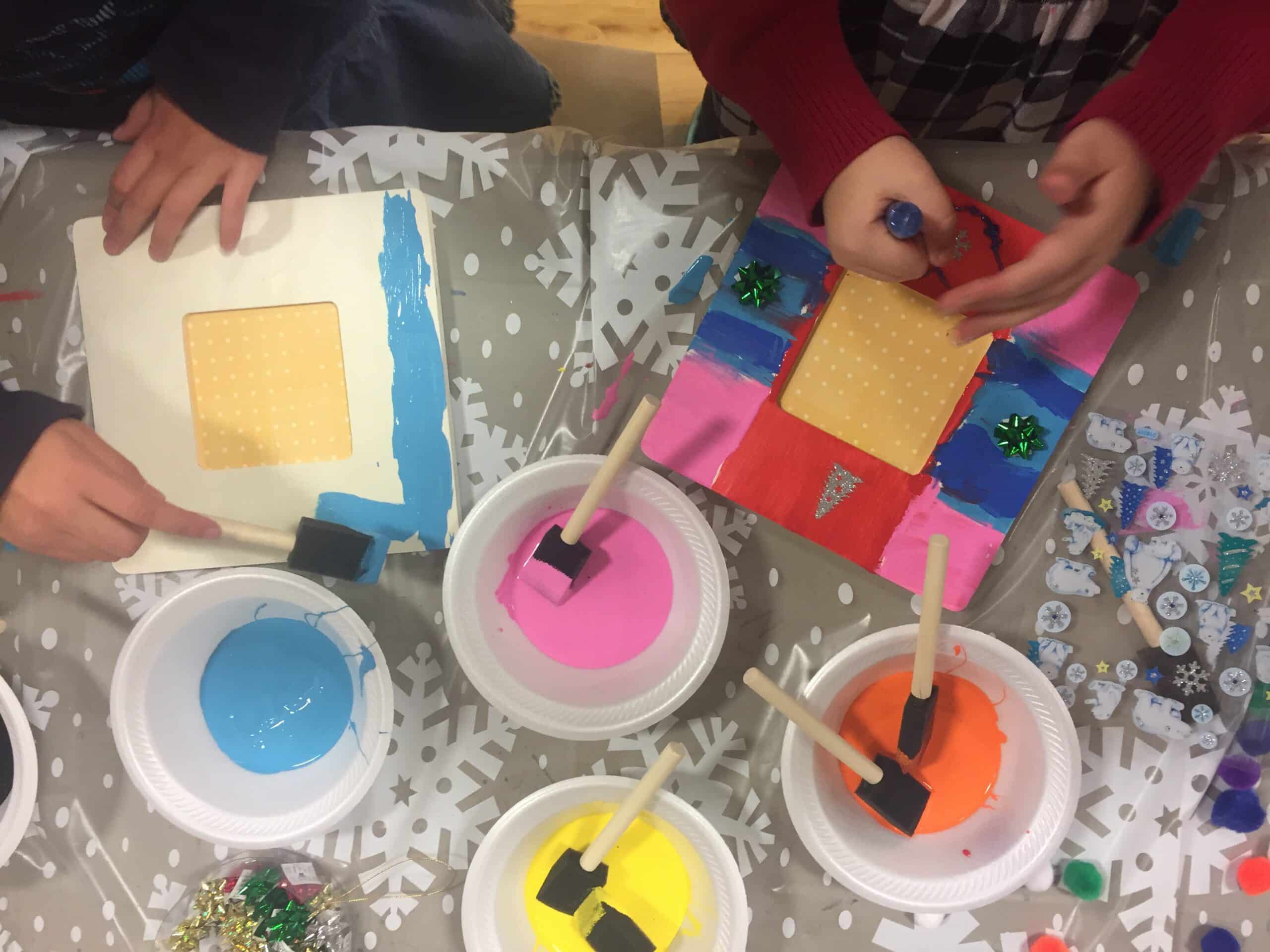 Overhead shot depicts children painting picture frames using foam brushes and multiple colors of paint. The picture frames are being painted with vibrant shades of blue, pink, and yellow. The mood is playful. This eye-level shot captures a glimpse into a creative activity, perfect for illustrating themes of education, arts and crafts, and childhood exploration. The photo would be appropriate for projects that celebrate art, family, learning, and creative expression and mindful makers