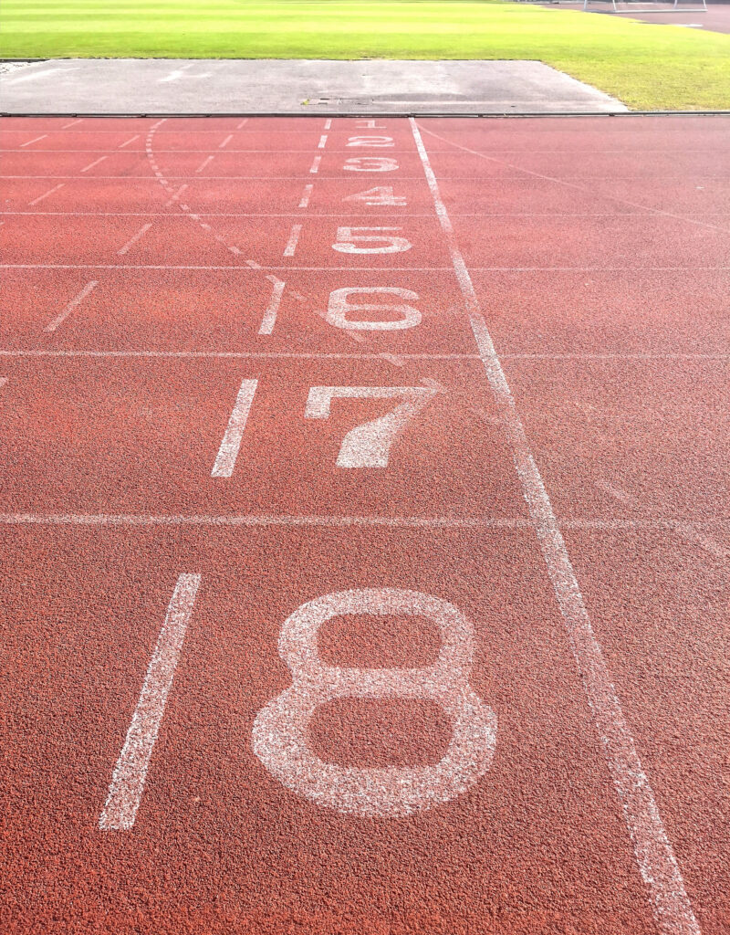 A striking image presents a ground-level view of an outdoor running track, highlighting lanes marked with numbers. The composition, set in bright daylight, draws attention to the textured red track surface and the clear, white lane markings. The photograph's focus and neutral color palette make it suitable for projects related to sports, fitness, competition, and urban environments. It is perfect for use in advertising material, editorial content, and educational resources.