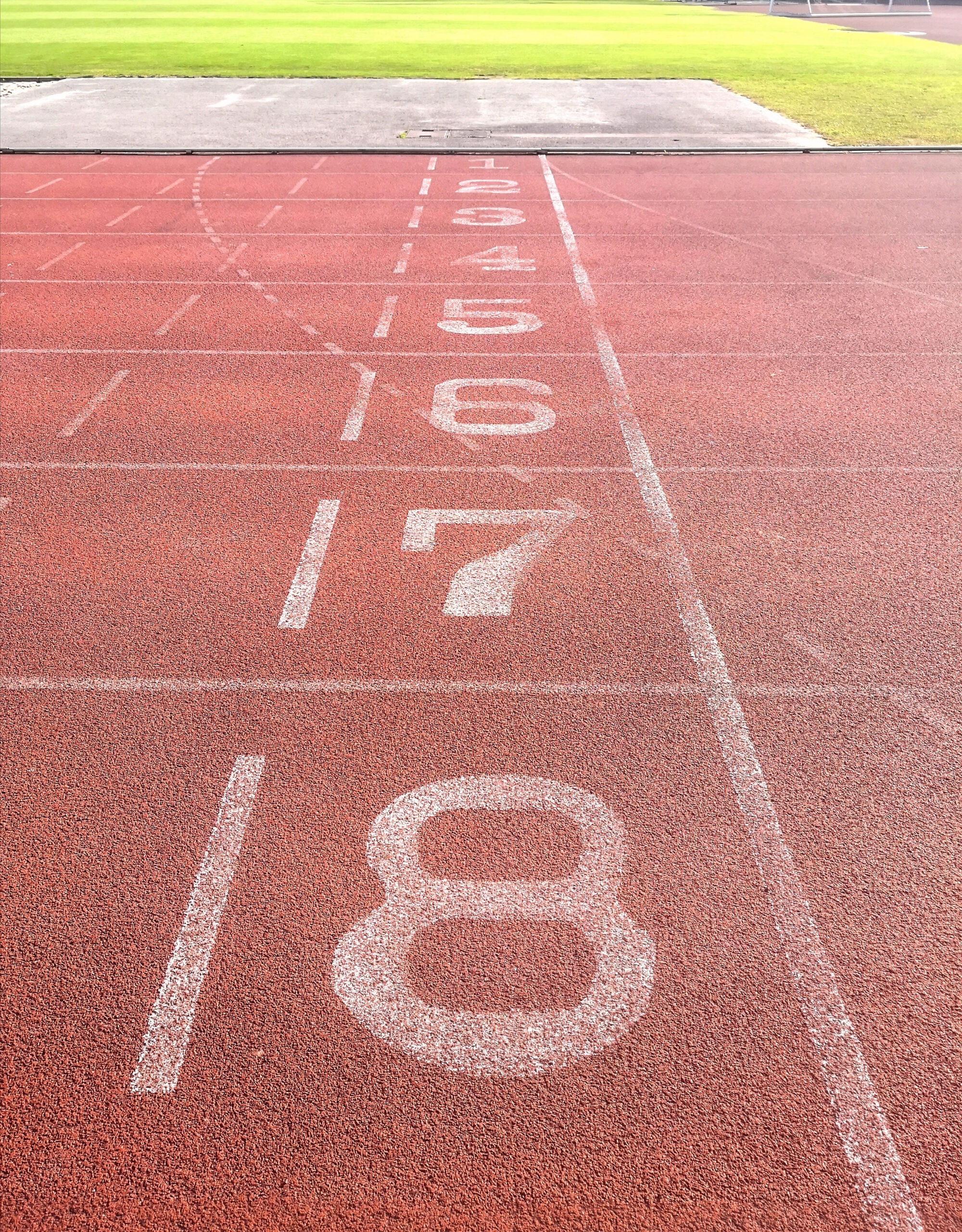 A striking image presents a ground-level view of an outdoor running track, highlighting lanes marked with numbers. The composition, set in bright daylight, draws attention to the textured red track surface and the clear, white lane markings. The photograph's focus and neutral color palette make it suitable for projects related to sports, fitness, competition, and urban environments. It is perfect for use in advertising material, editorial content, and educational resources.