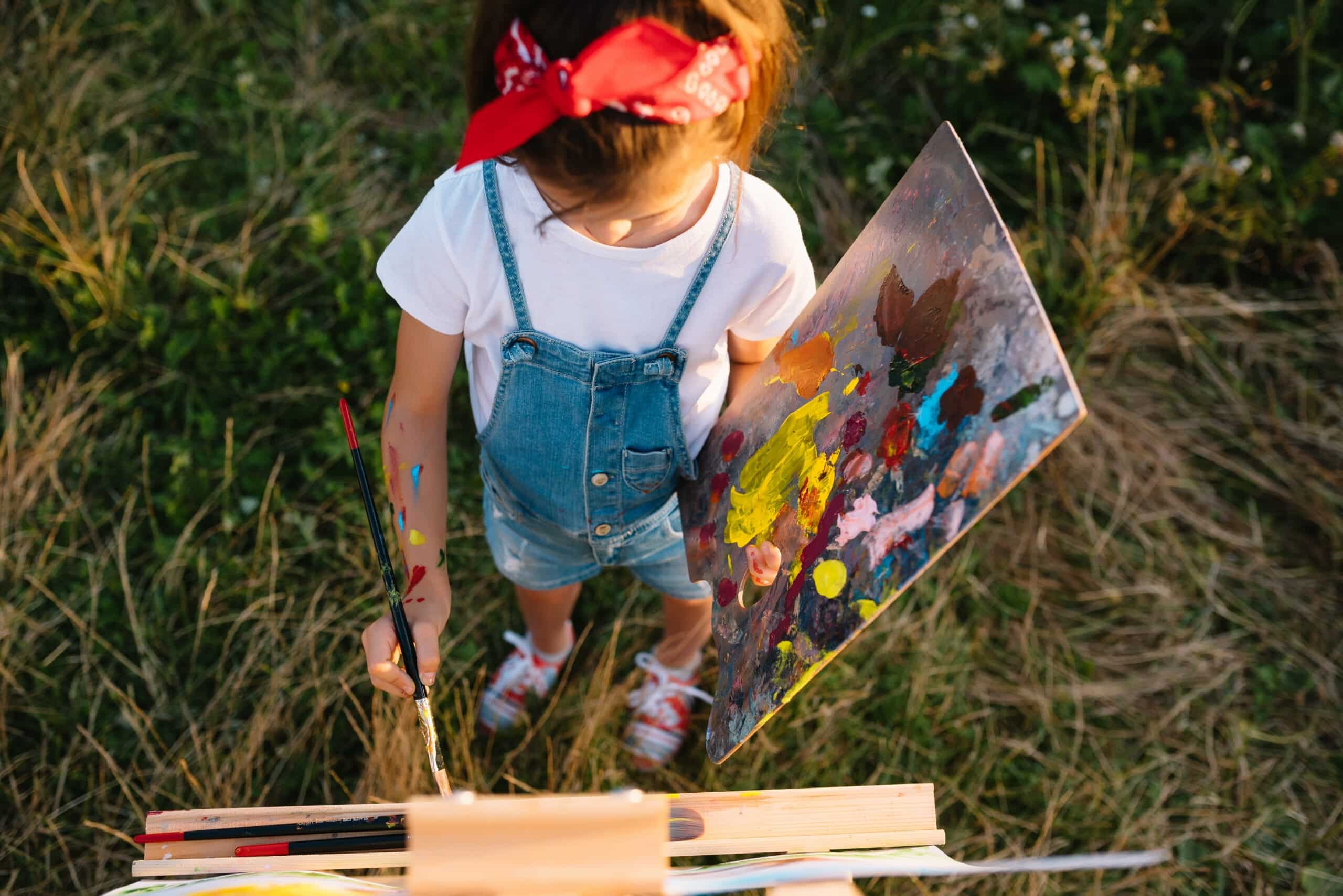 A child with light brown hair and a red bandana is outside in a grassy field painting on a canvas. The child is wearing a white t-shirt and blue jean overalls. The artwork and the child's arm have paint smeared on them. The photo shows a playful moment of a child in nature creating art. This image is suitable for projects about creativity, learning, childhood, and nature
