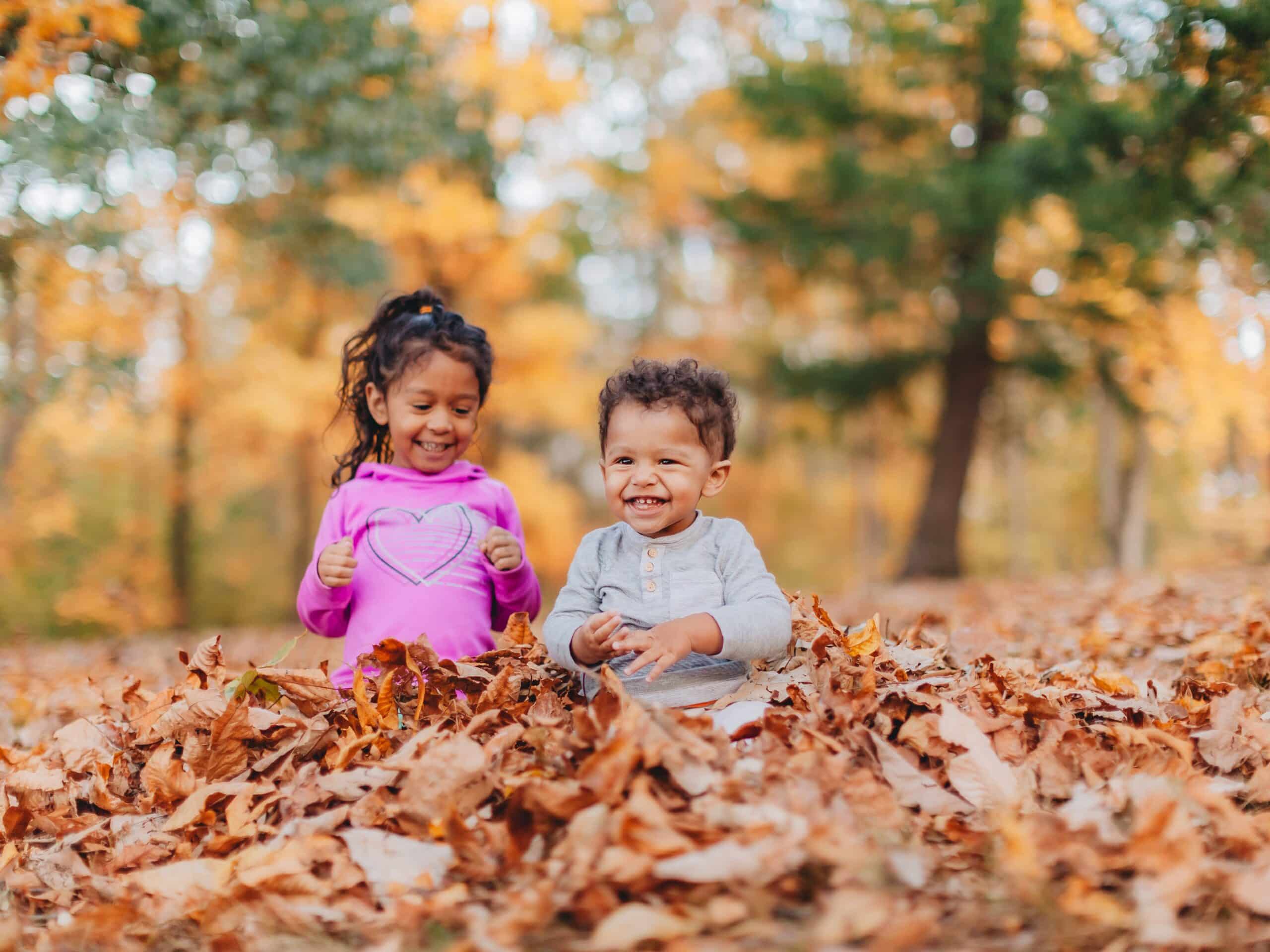 A curly-haired toddler and a child with long dark hair are smiling and playing together in a pile of colorful autumn leaves. The children are framed by a beautiful blurred forest in the background with trees showing yellow and green foliage. The soft lighting and the playful mood create a joyful, light-hearted scene, perfect for projects centered around family, autumn, childhood, or seasonal themes. The photo is an idyllic depiction of childhood joy and outdoor play